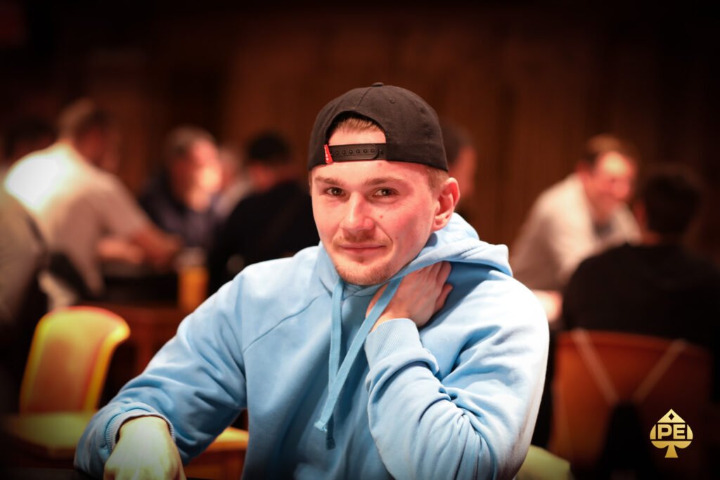 Ein Mann sitzt an einem Pokertisch in einem eleganten Casino, umgeben von anderen Spielern. Er hält Karten in der Hand und konzentriert sich auf das Spiel. Die Atmosphäre ist gespannt und glamour