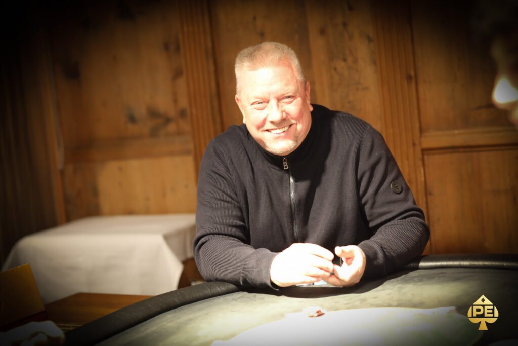 Ein professioneller Pokertisch mit grünem Filz, umgeben von Stühlen und beleuchtet von einer eleganten Lampe über dem Tisch. Karten und Chips sind auf dem Tisch verteilt, während Spieler in an