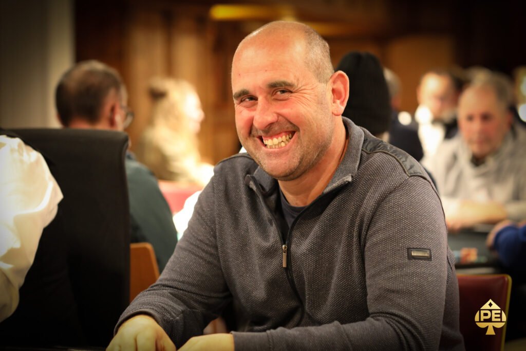 Ein Mann in einem dunkelblauen Anzug und einer Krawatte sitzt an einem Pokertisch und hält Pokerkarten in der Hand, während er konzentriert auf seine Gegner schaut. Die Szene spielt