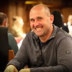 Ein Mann in einem dunkelblauen Anzug und einer Krawatte sitzt an einem Pokertisch und hält Pokerkarten in der Hand, während er konzentriert auf seine Gegner schaut. Die Szene spielt