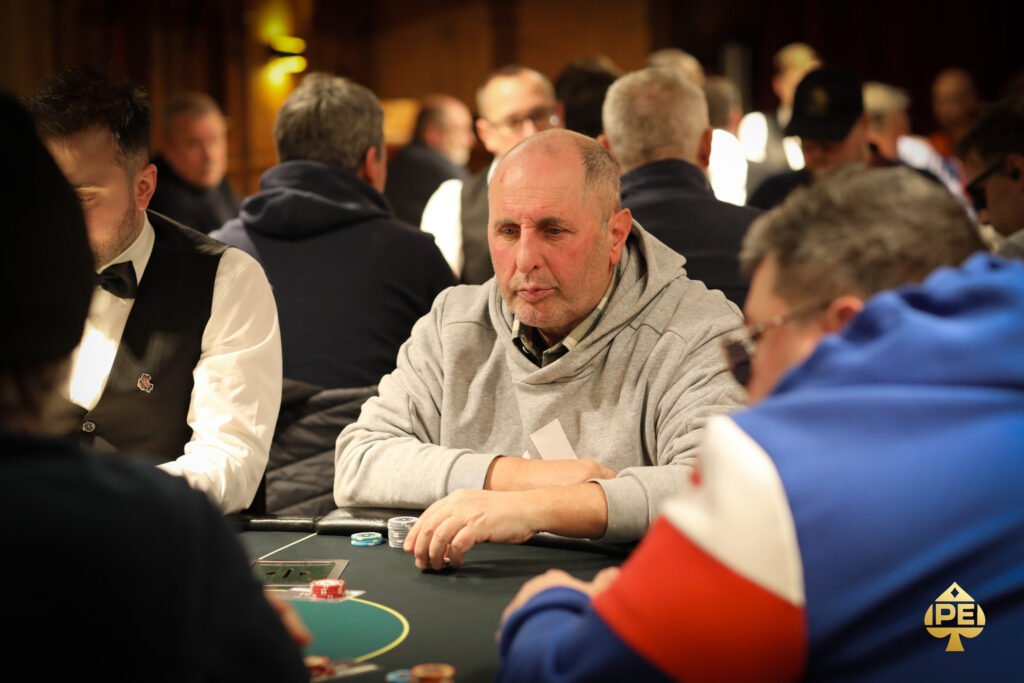 Ein Mann sitzt an einem Pokertisch und hält seine Karten in der Hand, während er konzentriert auf die Mitte des Tisches schaut. Andere Spieler sind im Hintergrund zu sehen.