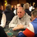 Ein Mann sitzt an einem Pokertisch und hält seine Karten in der Hand, während er konzentriert auf die Mitte des Tisches schaut. Andere Spieler sind im Hintergrund zu sehen.