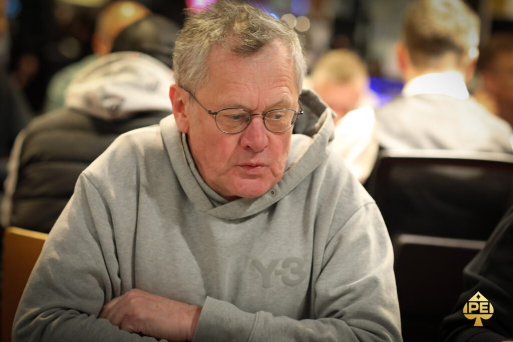 Ein Mann in einem dunkelblauen Anzug und einer weißen Hemdweste sitzt an einem Pokertisch und hält zwei Spielkarten in der Hand. Er hat einen ernsten Gesichtsausdruck und schaut