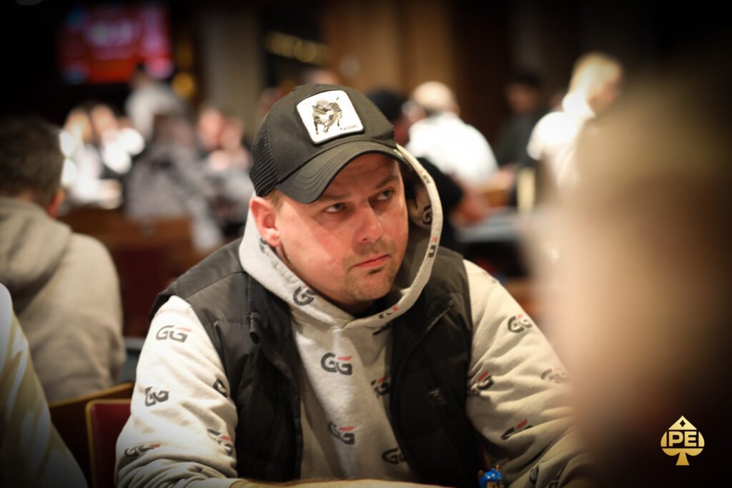 Ein professioneller Pokerspieler mit Sonnenbrille und Kappe sitzt an einem Spieltisch und betrachtet konzentriert seine Karten. Um ihn herum sind Chips und Karten verstreut.