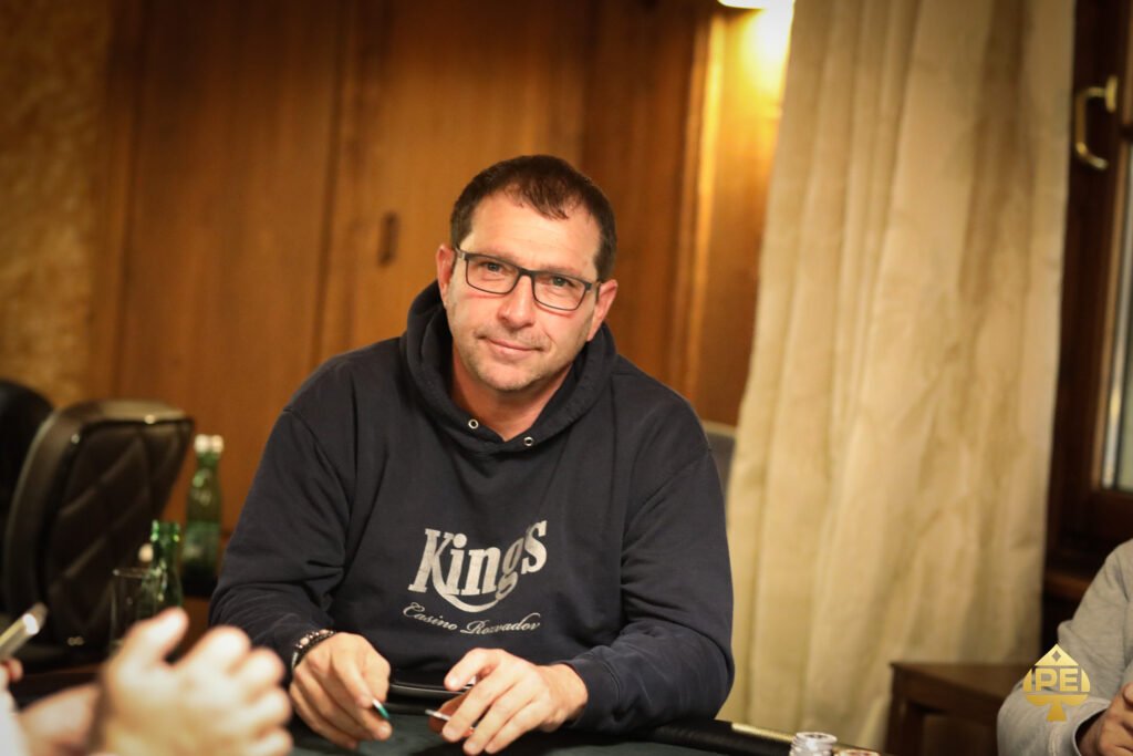 Ein professioneller Pokerspieler in einem dunklen Anzug sitzt an einem Pokertisch und betrachtet konzentriert seine Karten. Im Hintergrund sind andere Spieler und Chips zu sehen.