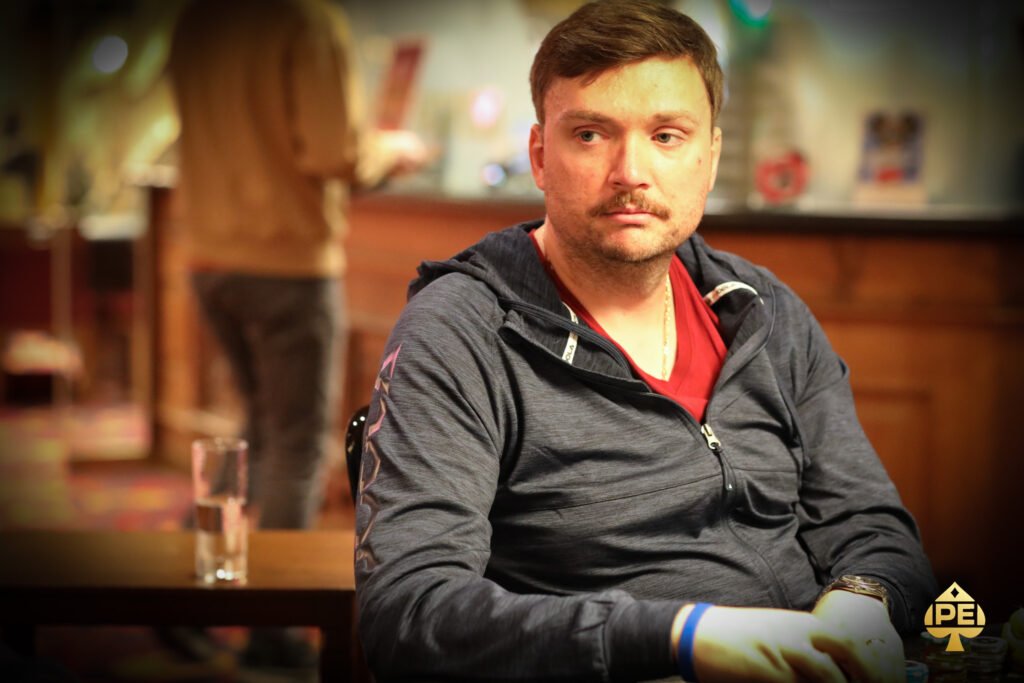 Ein Mann in einem dunkelblauen Anzug und einer Krawatte spielt konzentriert Poker an einem grünen Pokertisch in einem eleganten Casino. Er hält Karten in der Hand und betrachtet mit ern