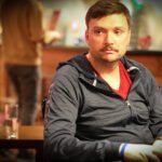Ein Mann in einem dunkelblauen Anzug und einer Krawatte spielt konzentriert Poker an einem grünen Pokertisch in einem eleganten Casino. Er hält Karten in der Hand und betrachtet mit ern