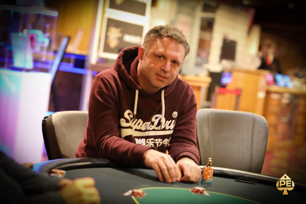 Ein professioneller Pokerspieler mit Sonnenbrille und Kappe sitzt an einem Pokertisch und betrachtet konzentriert seine Karten. Im Hintergrund sind weitere Spieler und Chips zu sehen.