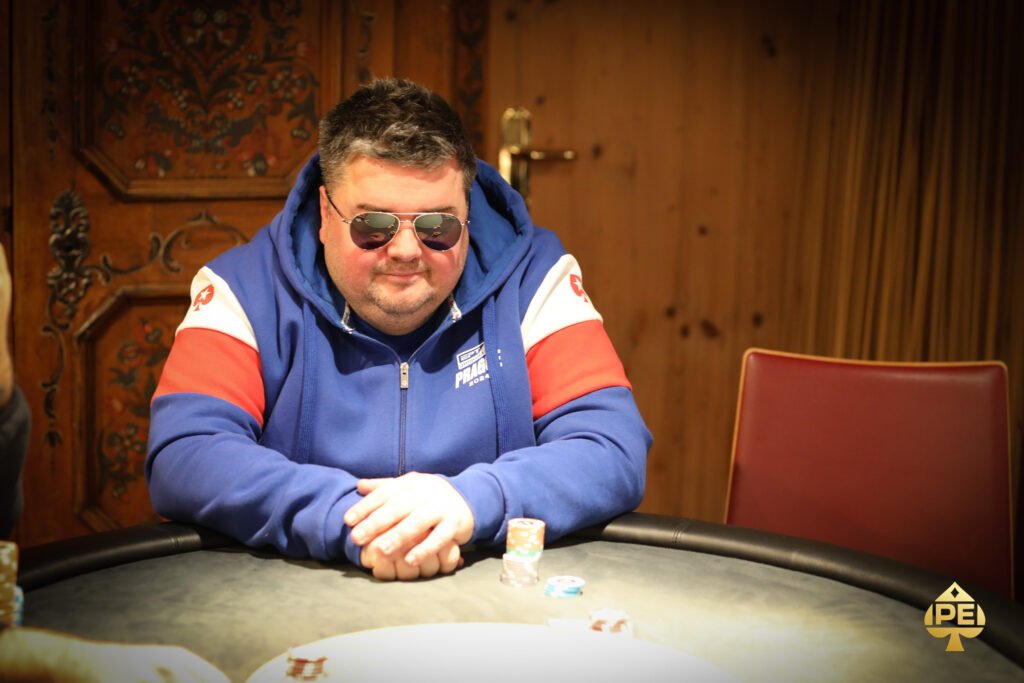 Ein Mann in einem dunkelblauen Anzug und einer Krawatte steht vor einer Pokertisch mit Spielkarten und Pokerchips. Er hält eine Hand mit Karten und schaut mit einem ernsten Gesichtsausdr