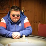 Ein Mann in einem dunkelblauen Anzug und einer Krawatte steht vor einer Pokertisch mit Spielkarten und Pokerchips. Er hält eine Hand mit Karten und schaut mit einem ernsten Gesichtsausdr