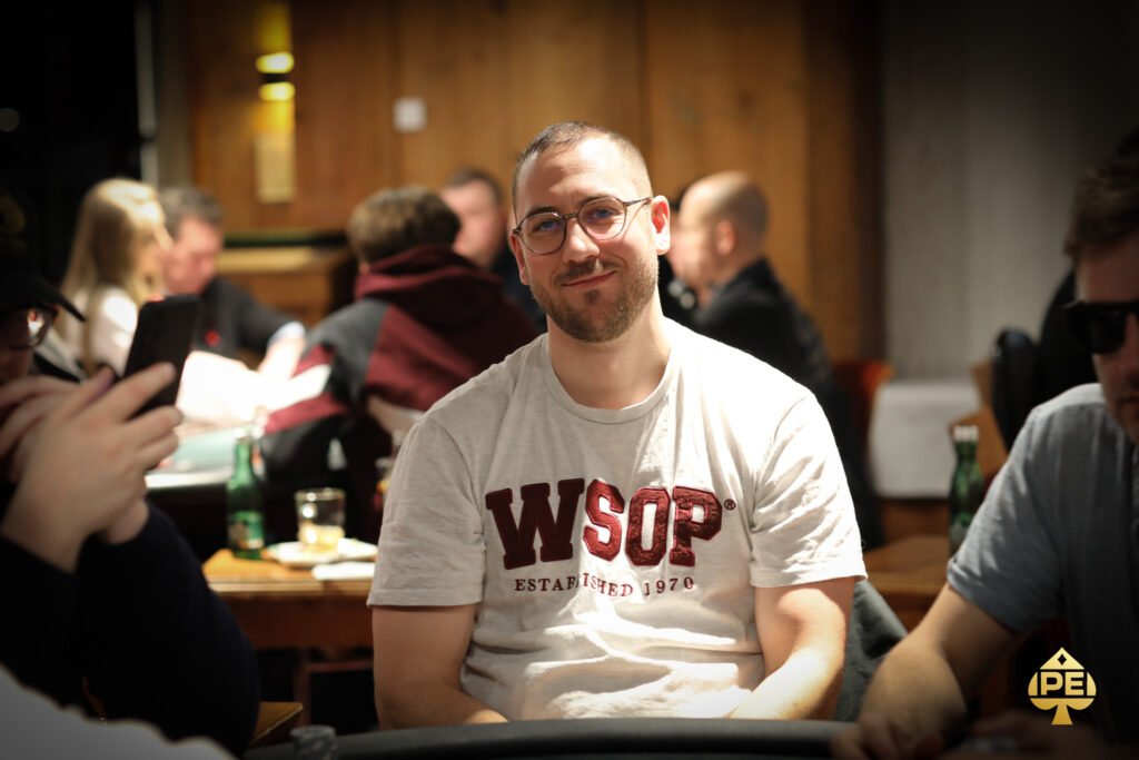 Ein Mann in einem dunkelblauen Anzug und einer roten Krawatte sitzt an einem Pokertisch und betrachtet konzentriert seine Karten. In der Mitte des Tisches liegen Chips und Spielkarten