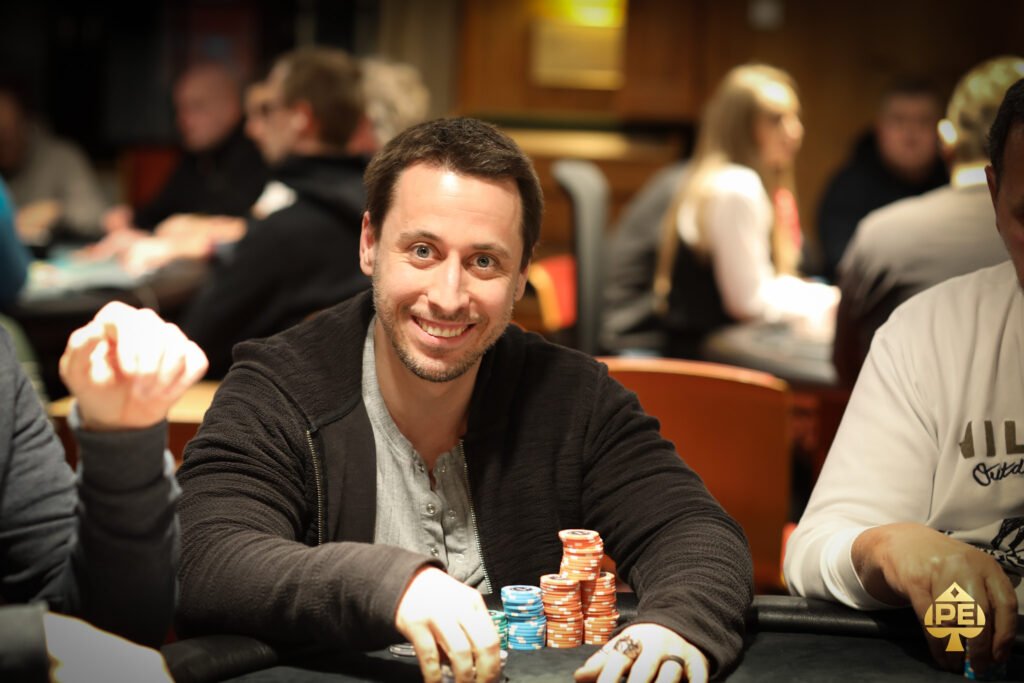 Ein Mann sitzt an einem Pokertisch und betrachtet konzentriert seine Karten. In der Hand hält er Pokerchips und neben ihm liegen Spielkarten auf dem Tisch. Im Hintergrund sind weitere Spieler
