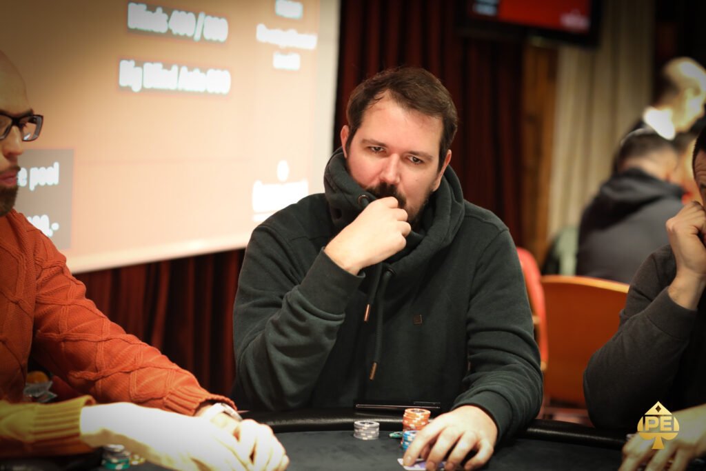 Ein professioneller Pokertisch mit grünem Filz, umgeben von Stühlen und Chips. Drei Spieler sitzen mit ernstem Gesichtsausdruck an ihren Plätzen, während sie Karten halten und auf ihre