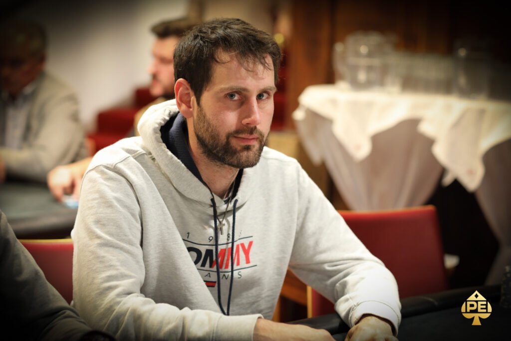 Ein Mann in dunkler Kleidung sitzt an einem Pokertisch und hält eine Hand mit Spielkarten. Er schaut konzentriert auf seine Karten, während im Hintergrund andere Spieler und Pokerchips zu sehen