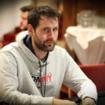 Ein Mann in dunkler Kleidung sitzt an einem Pokertisch und hält eine Hand mit Spielkarten. Er schaut konzentriert auf seine Karten, während im Hintergrund andere Spieler und Pokerchips zu sehen