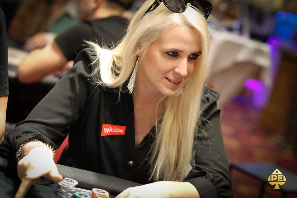 Ein Mann sitzt an einem Pokertisch und hält ein Paar Asse in der Hand. Er trägt eine schwarze Sonnenbrille und konzentriert sich auf seine Karten. Andere Spieler sind im Hintergrund