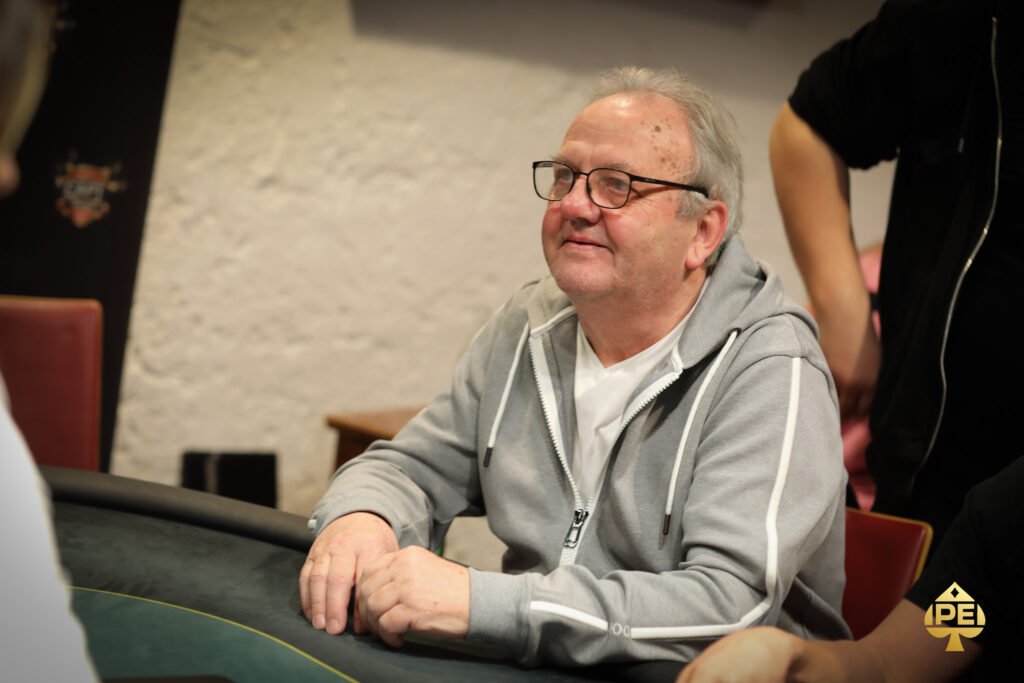 Ein Mann in einem schwarzen Anzug und einer blauen Krawatte sitzt an einem Pokertisch und betrachtet konzentriert seine Karten. Im Hintergrund sind weitere Spieler und Casinolichter zu sehen.
