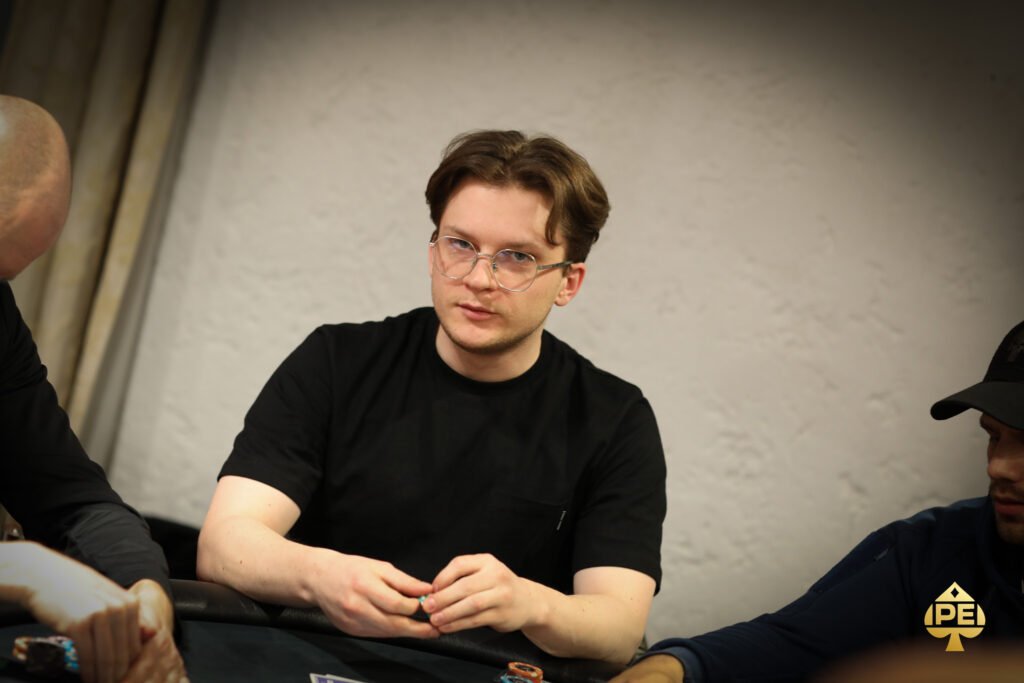 Ein Mann sitzt an einem Pokertisch und hält seine Karten. Er trägt ein schwarzes Hemd und eine schwarze Kappe. Auf dem Tisch liegen Pokerchips und Karten. Im Hintergrund sind weitere