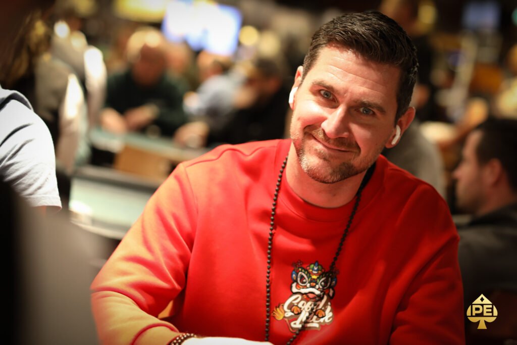 Ein Mann sitzt an einem Pokertisch mit grünem Filz und hält zwei Spielkarten in der Hand. Er trägt eine schwarze Kappe und konzentriert sich auf das Spiel. Im Hintergrund sind Poker
