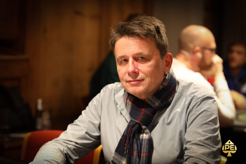 Ein Mann sitzt an einem Pokertisch, umgeben von anderen Spielern. Er hält seine Karten in der Hand und konzentriert sich auf das Spiel. Die Atmosphäre ist gespannt und fokussiert.