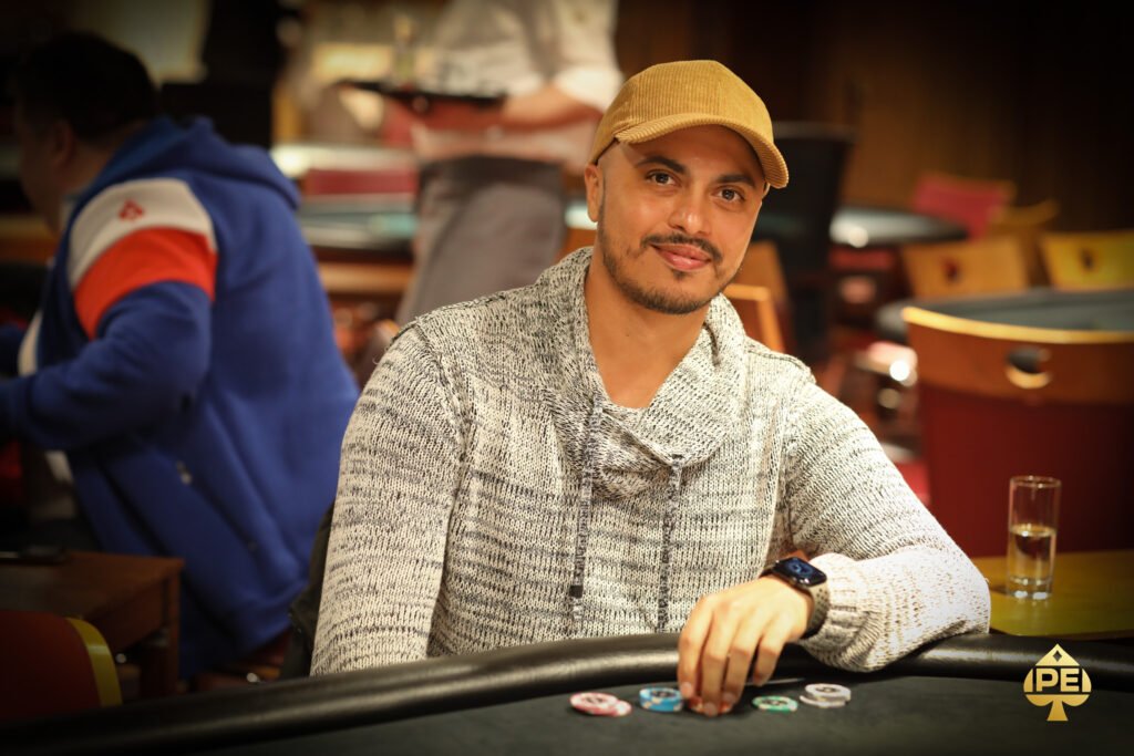 Ein Mann in einem schwarzen Anzug und einer schwarzen Sonnenbrille sitzt an einem Pokertisch und hält eine Hand mit Spielkarten. In der Mitte des Tisches liegen weitere Karten und Pokerchips.