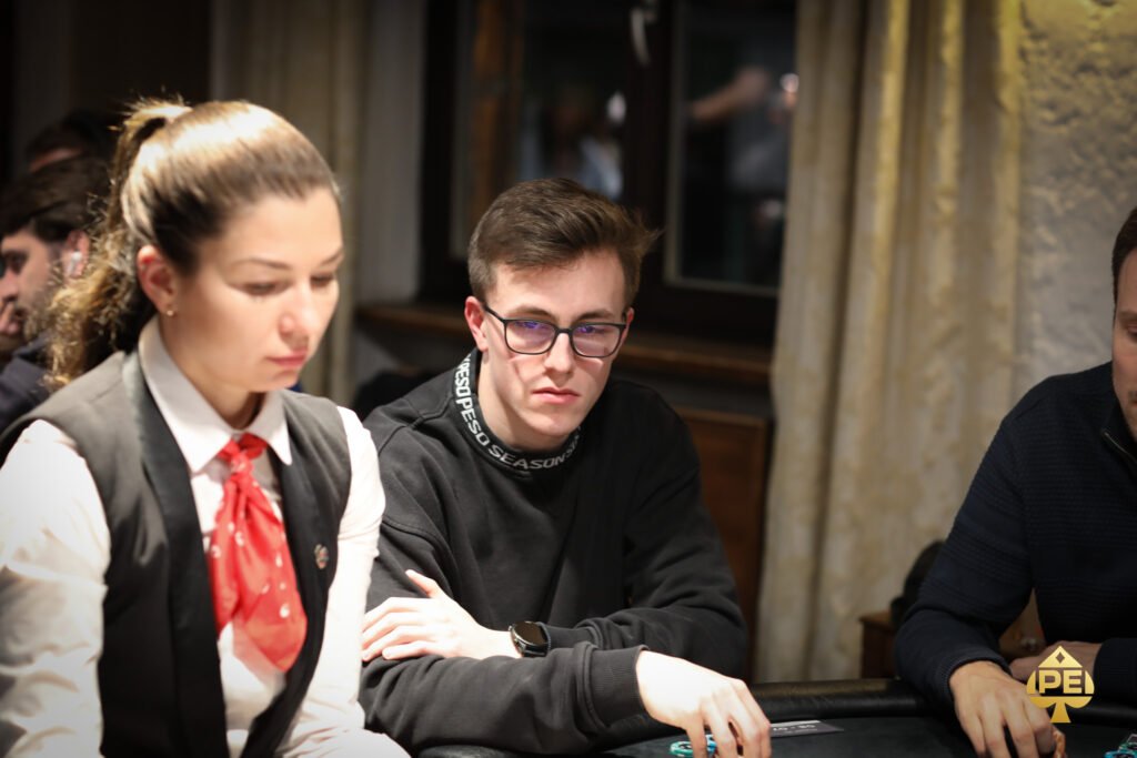 Ein Mann in einem dunkelblauen Anzug sitzt an einem Pokertisch und hält zwei Spielkarten in der Hand. Er schaut konzentriert auf den Tisch, auf dem Pokerchips und Spielkarten verte