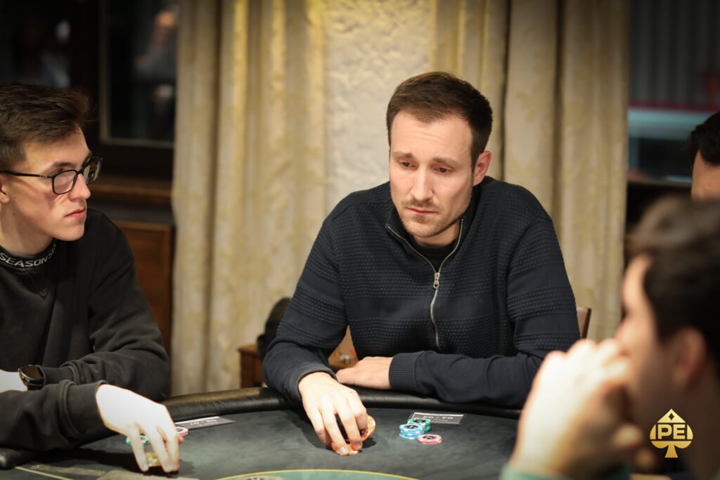 Ein Mann in einem Anzug und mit einer Sonnenbrille spielt Poker an einem Tisch in einem luxuriösen Casino. Er hält Karten in der Hand und schaut konzentriert auf den Tisch. Andere