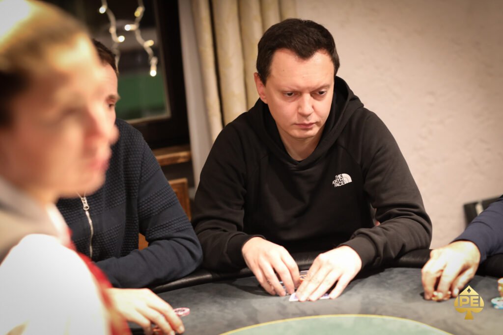 Ein Mann in einem schwarzen Anzug und weißem Hemd sitzt an einem Pokertisch und hält zwei Spielkarten in der Hand. Er schaut konzentriert auf den Tisch, auf dem Pokerchips und K