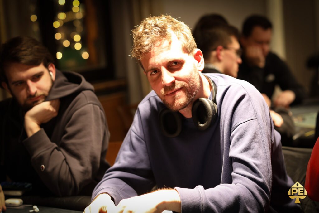 Ein Mann in einem dunklen Anzug sitzt an einem Pokertisch und hält Karten in der Hand. Im Hintergrund sind andere Spieler und Pokerchips zu sehen.