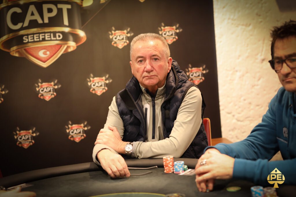 Ein Mann sitzt an einem Pokertisch und betrachtet konzentriert seine Karten. Im Hintergrund sind andere Spieler und Pokerchips zu sehen.