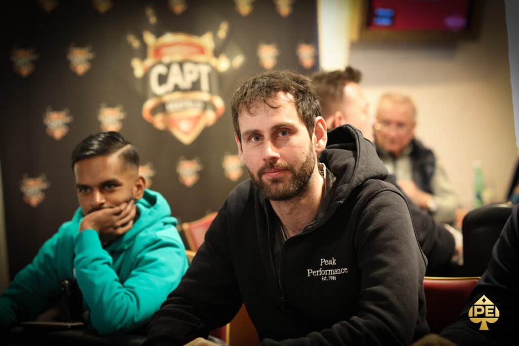 Ein Mann in einem schwarzen Anzug und einer Fliege sitzt an einem Pokertisch und hält eine Hand mit Spielkarten, während er konzentriert in die Kamera schaut. Im Hintergrund sind Pokerch