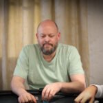 Ein Mann und eine Frau sitzen an einem Pokertisch im Casino und spielen konzentriert. Die Frau hält Karten in der Hand und der Mann schaut auf seine Chips. Im Hintergrund sind andere Spieler und Dealer zu sehen