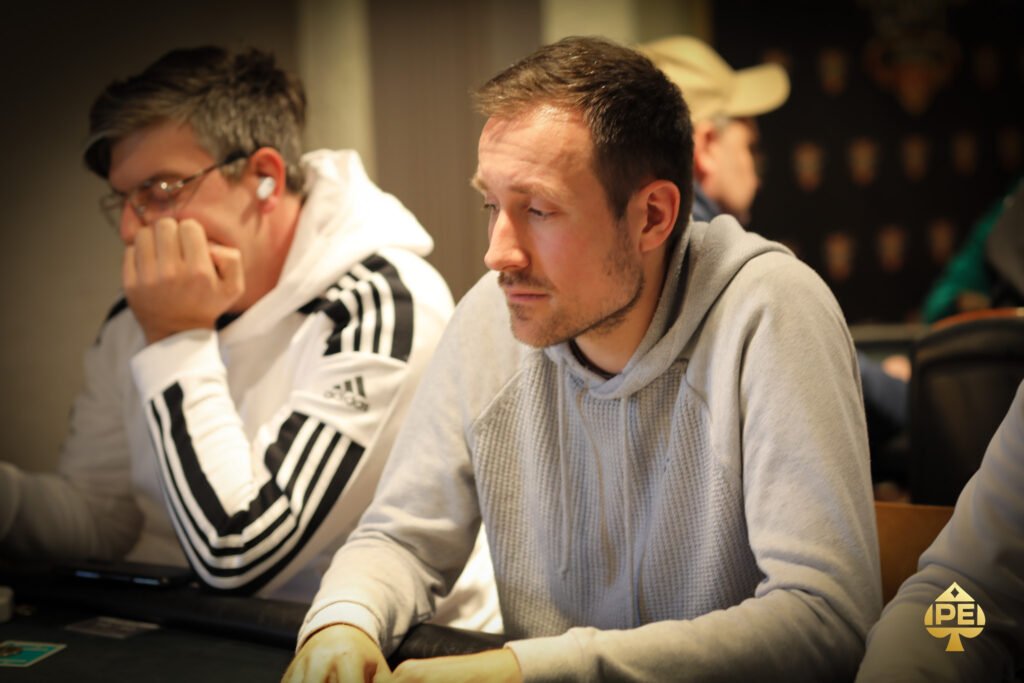 Ein Mann und eine Frau sitzen an einem Pokertisch und spielen gegeneinander. Der Mann hat eine Pokerhand und schaut konzentriert auf seine Karten, während die Frau mit einem selbstbewussten Blick auf ihre Chips
