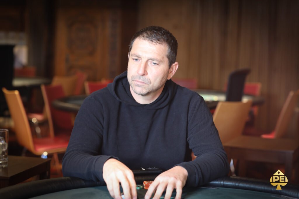 Ein Mann sitzt an einem Pokertisch und hält seine Karten in der Hand, während er konzentriert auf den Tisch schaut. Andere Spieler sind im Hintergrund zu sehen.