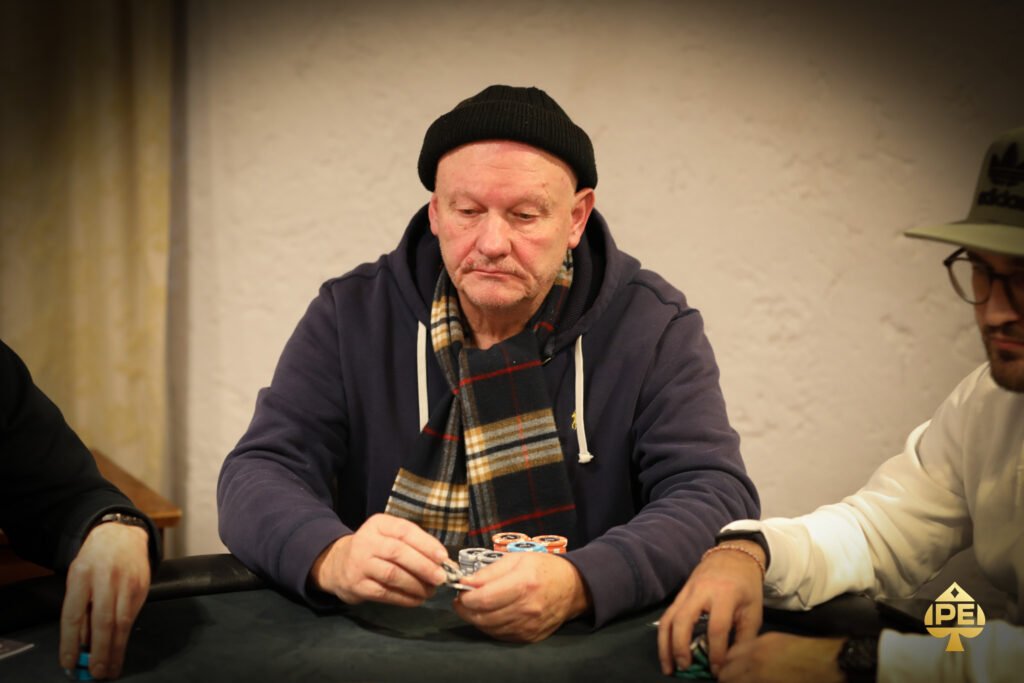 Ein Mann und eine Frau sitzen lächelnd an einem Pokertisch und halten Karten in der Hand, während sie sich in einem Casino befinden. Im Hintergrund sind weitere Spieler und Spieltische zu sehen.