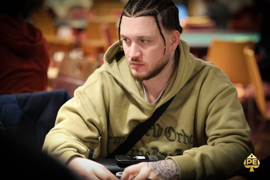 Ein Mann in einem dunklen Anzug steht vor einem Pokertisch mit Spielkarten und Pokerchips. Er hat eine ernste Miene und hält einen Stapel Chips in der Hand. Andere Spieler sind im Hintergrund zu