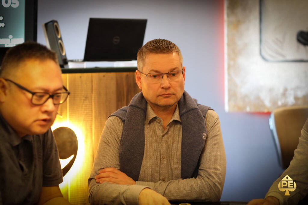 Ein Mann sitzt an einem Pokertisch und hält zwei Spielkarten in der Hand, während er konzentriert auf seine Gegner schaut. Im Hintergrund sind Pokerchips und Getränke zu sehen.