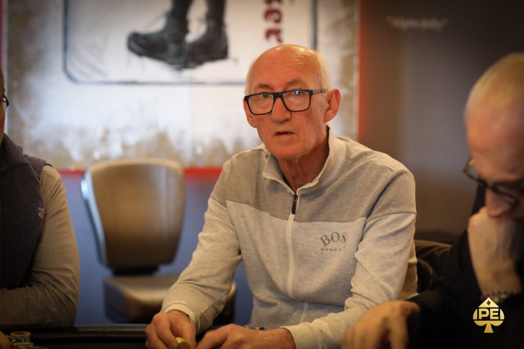 Ein Mann in einem dunkelblauen Anzug und einer roten Krawatte sitzt an einem Pokertisch und betrachtet konzentriert seine Karten. Im Hintergrund sind verschwommene Personen und Casino-Licht