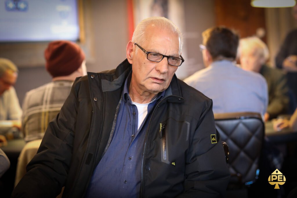 Ein Mann in einem schwarzen Anzug und einer schwarzen Sonnenbrille sitzt an einem Pokertisch und hält zwei Spielkarten in der Hand. Er wirkt konzentriert und ernst. Im Hintergrund sind