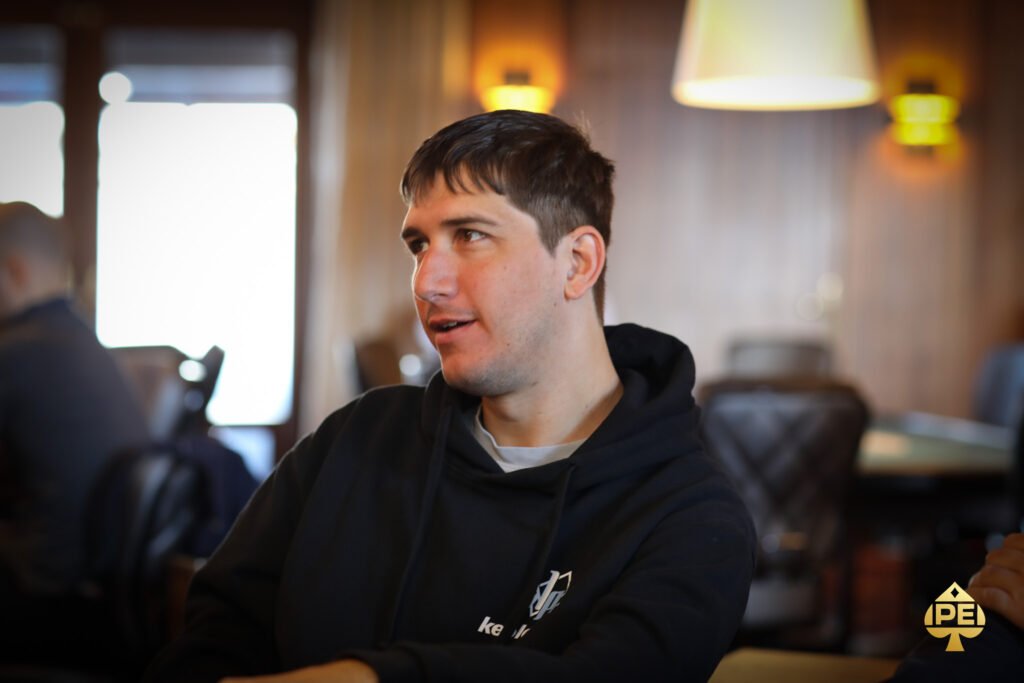 Ein Mann sitzt an einem Pokertisch und hält zwei Spielkarten in der Hand. Er schaut konzentriert auf den Tisch, wo Pokerchips und weitere Karten liegen. Im Hintergrund sind weitere Spieler