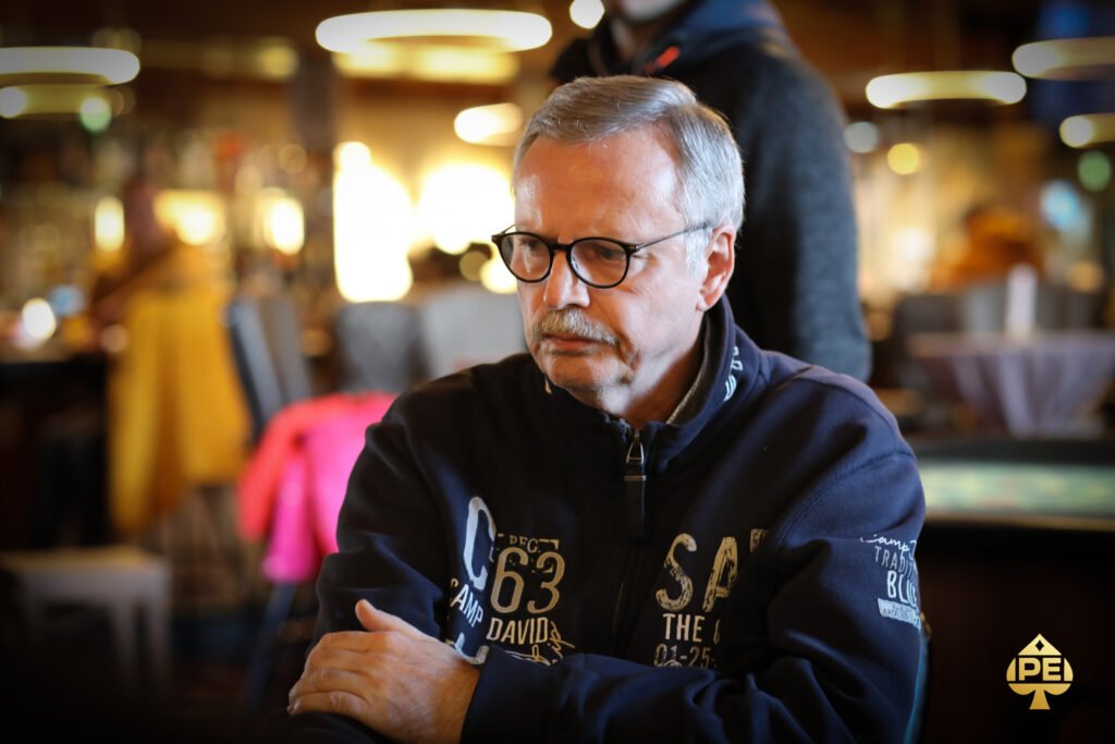 Ein Spieler, der eine Pokerhand hält und seine Karten betrachtet, während er an einem Pokertisch sitzt. Andere Spieler und Chips sind im Hintergrund zu sehen.