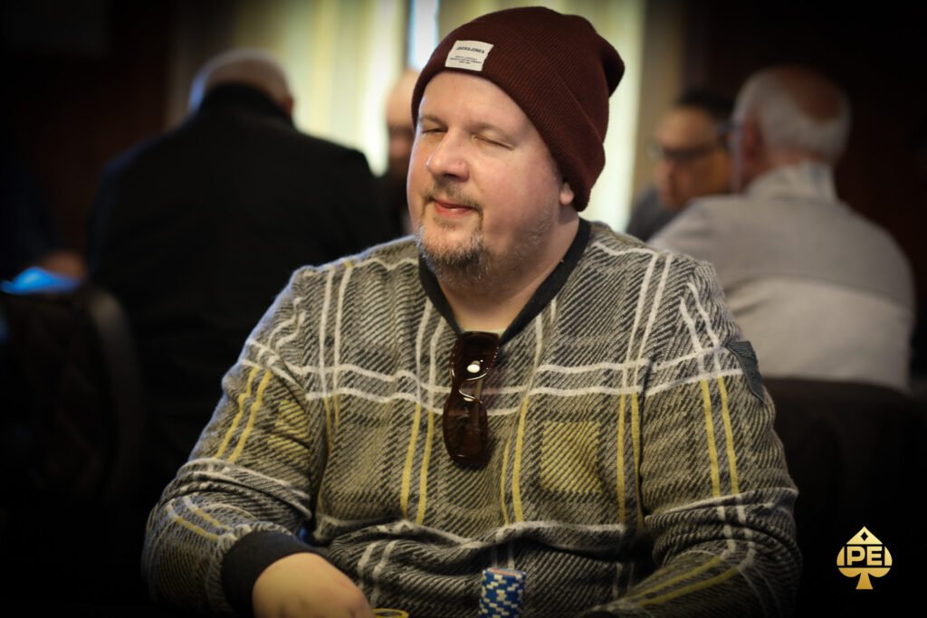 Ein Mann sitzt an einem Pokertisch und hält zwei Spielkarten in der Hand, während er konzentriert auf die Gemeinschaftskarten in der Mitte des Tisches schaut. Im Hintergrund sind weitere Spieler