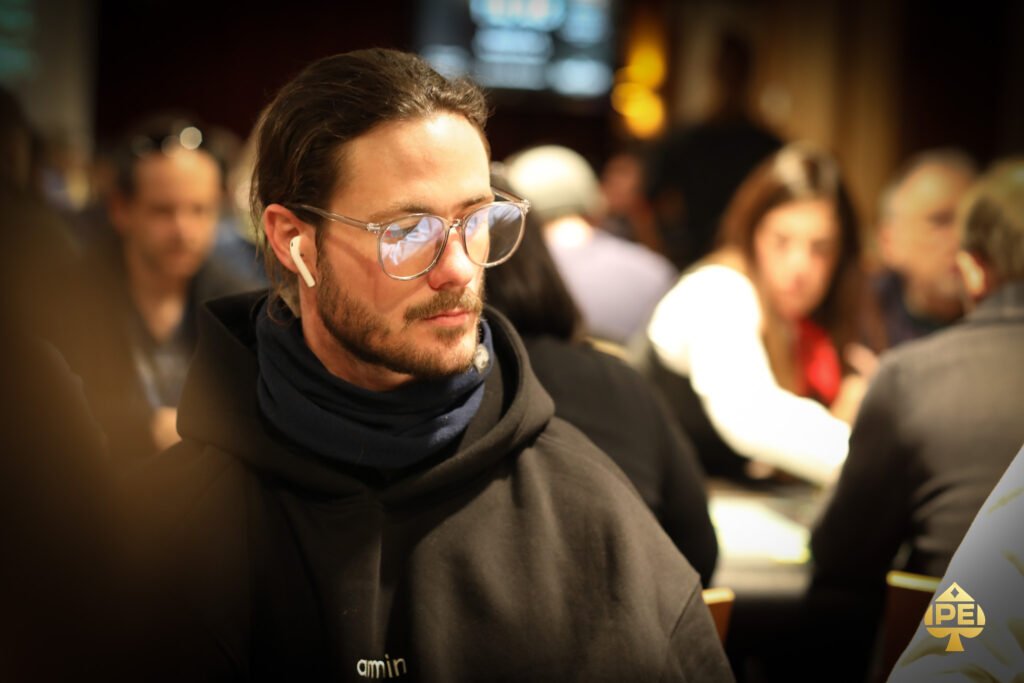 Ein Mann in einem schwarzen Anzug und einer blauen Krawatte spielt im Casino an einem Pokertisch. Er hält Karten in der Hand und schaut konzentriert auf seine Gegner. Die Atmosphäre ist