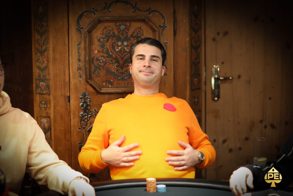 Ein professioneller Pokerspieler mit Sonnenbrille und Kappe sitzt an einem Pokertisch und betrachtet seine Karten mit ernstem Gesichtsausdruck. Im Hintergrund sind andere Spieler und Jetons zu