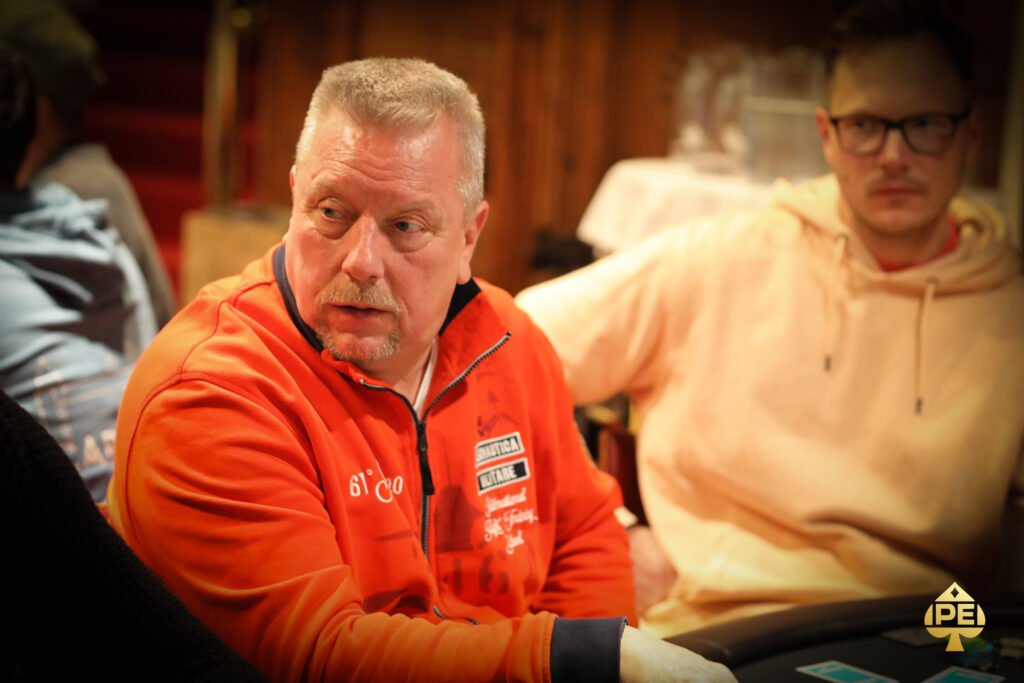 Ein Mann und eine Frau sitzen an einem Pokertisch und spielen eine Partie Poker. Der Mann hält Karten in der Hand, während die Frau konzentriert auf ihre eigenen Karten schaut. Im Hintergrund sind Poker