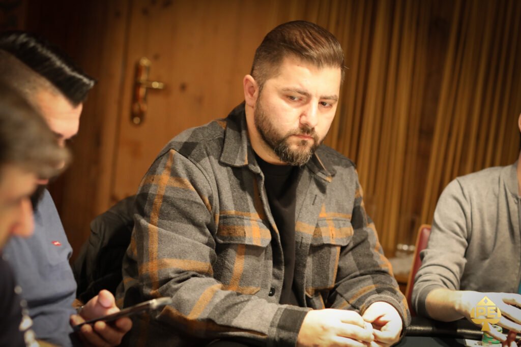 Ein Mann sitzt an einem Pokertisch und betrachtet konzentriert seine Karten. Im Hintergrund sind andere Spieler und Pokerchips zu sehen.