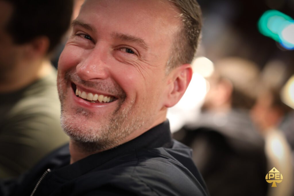 Ein Mann sitzt an einem Pokertisch und hält seine Karten in der Hand, während er konzentriert auf den Spielverlauf schaut. Im Hintergrund sind weitere Spieler und Chips zu sehen.