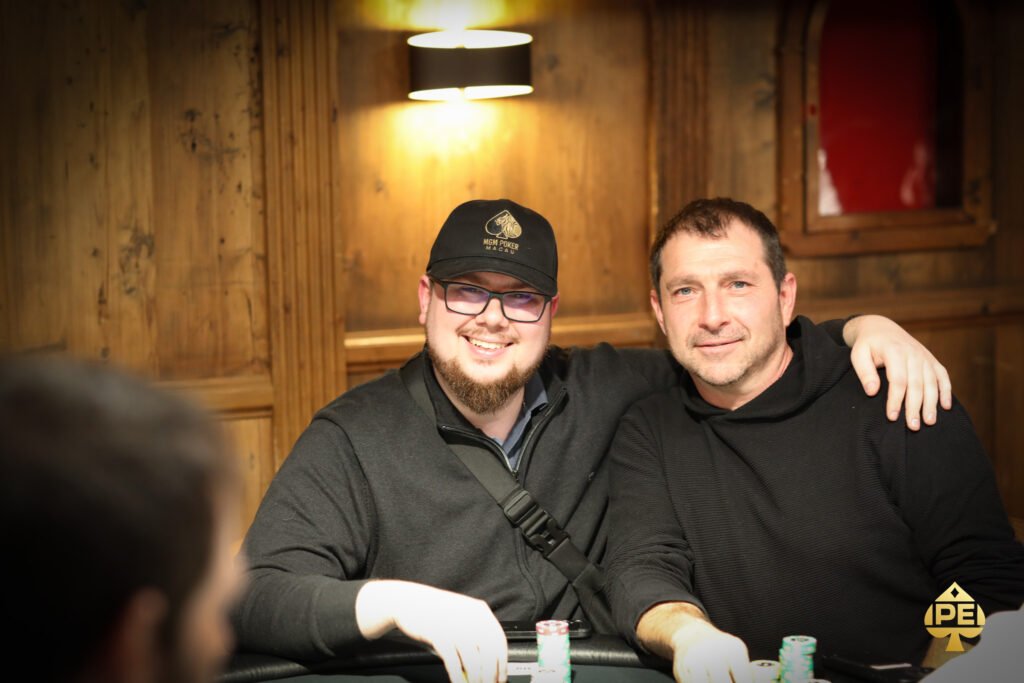 Ein Mann sitzt an einem Pokertisch und hält zwei Spielkarten in der Hand, während er konzentriert auf seine Gegner schaut. Die Szene spielt sich in einem eleganten Pokerraum mit grünen