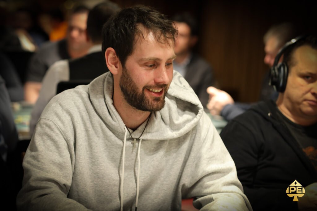 Ein Mann sitzt an einem Pokertisch und hält zwei Spielkarten in der Hand, während er konzentriert auf die Mitte des Tisches schaut. Andere Spieler sind im Hintergrund zu sehen.