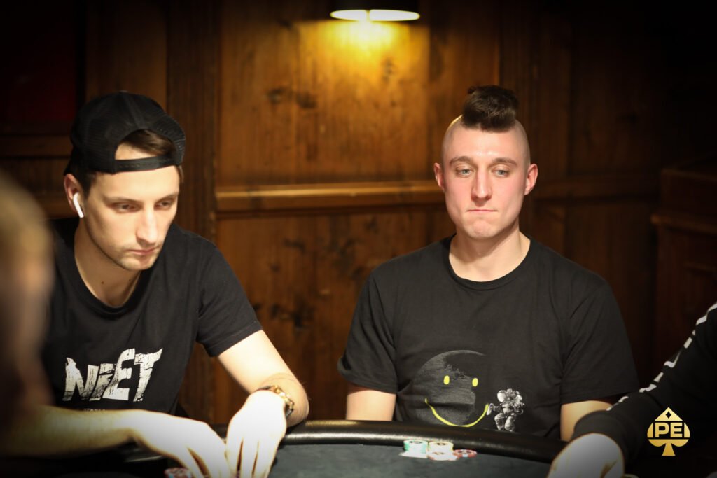 Ein Mann sitzt an einem Pokertisch und hält zwei Karten in der Hand. Er schaut konzentriert auf den Tisch, auf dem Pokerchips und Spielkarten liegen. Im Hintergrund sind andere Spieler